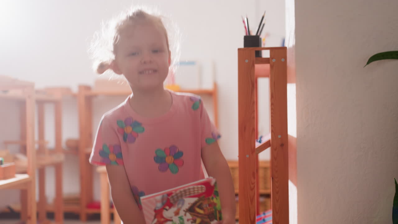 Toddler girl in kindergarten holding children story book waving goodbye with happy expression as sunlight streams through window, standing near wooden shelves in bright classroom environment