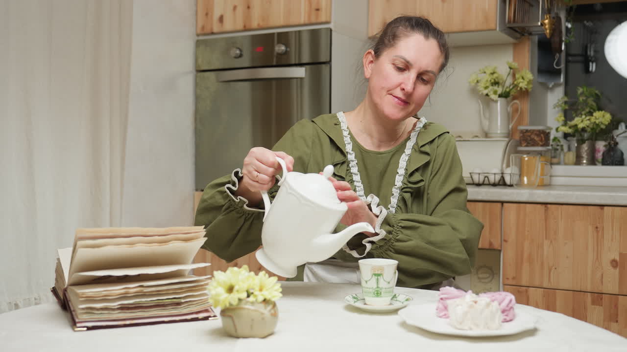 Woman in green dress gently pours herbal tea from white ceramic teapot into transparent glass cup while sitting at kitchen table with open recipe book, yellow flowers, and pastel cupcakes beside her