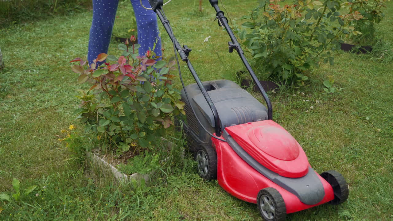 Young woman is mowing lawn with lawn mower in her back yard. Electric lawn mower cuts the grass.