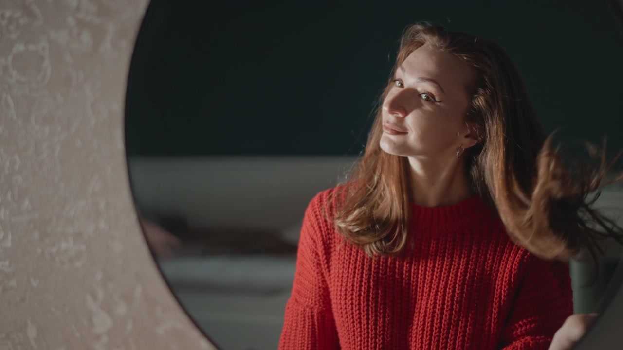 Long-haired woman wearing red knit sweater looks toward round wall mirror while gesturing, smoothing hair, and speaking in cozy bedroom setting with warm directional light, relaxed expression, and