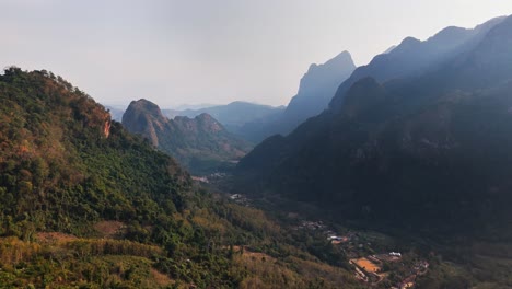 Drone captures dramatic limestone karst mountains of Nong Khiaw, Laos, as they rise above a winding forested valley scattered with rural farms and traditional villages in the golden afternoon light