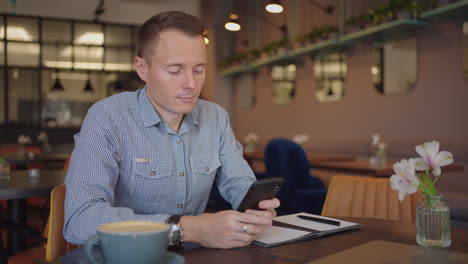 medium shot of young man wearing streetwear sitting in restaurant or cafe in front of laptop and having mobile phone. breakfast with smartphone