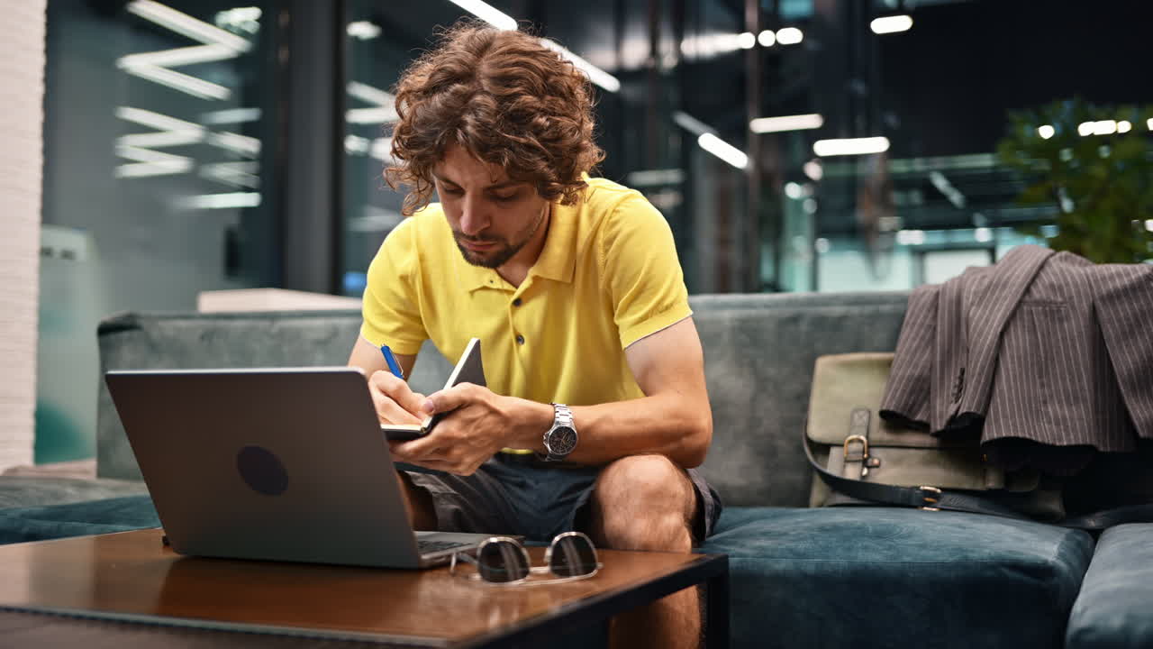 Man in yellow shirt standing on a couch and writing in a notebook and working on a computer