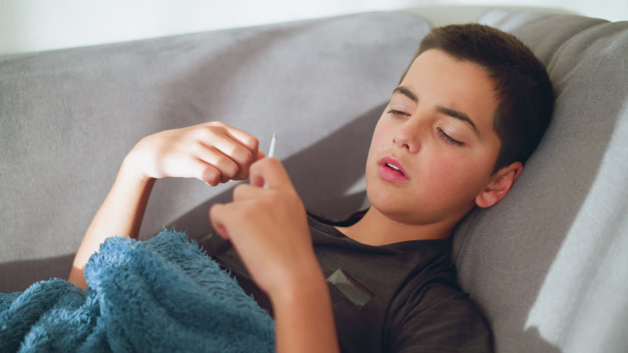 Boy collecting thermometer from someone to check his temperature, looking sick, resting with mouth slightly open, blue blanket covering him
