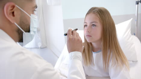 diverse male doctor in mask checking eyes of girl patient in hospital bed with penlight, slow motion