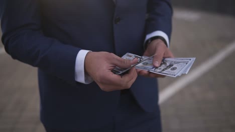 Solid-rich-businessman-on-the-street-holding-bundle-of-american-money-in-his-hands,-counting-it.-Wealthy-male-counts-hundred