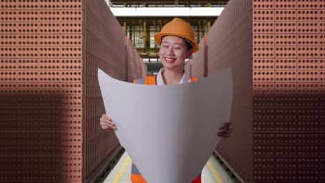 asian female engineer with safety helmet looking at blueprint in her hands while standing with red brick packed in stacks are stored