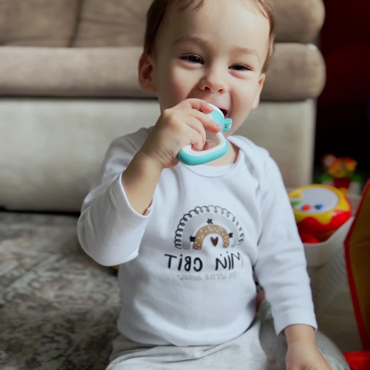 Happy smiling little Caucasian toddler sits on the floor. Baby in white suit eating a candy. Child active time at home. Vertical video