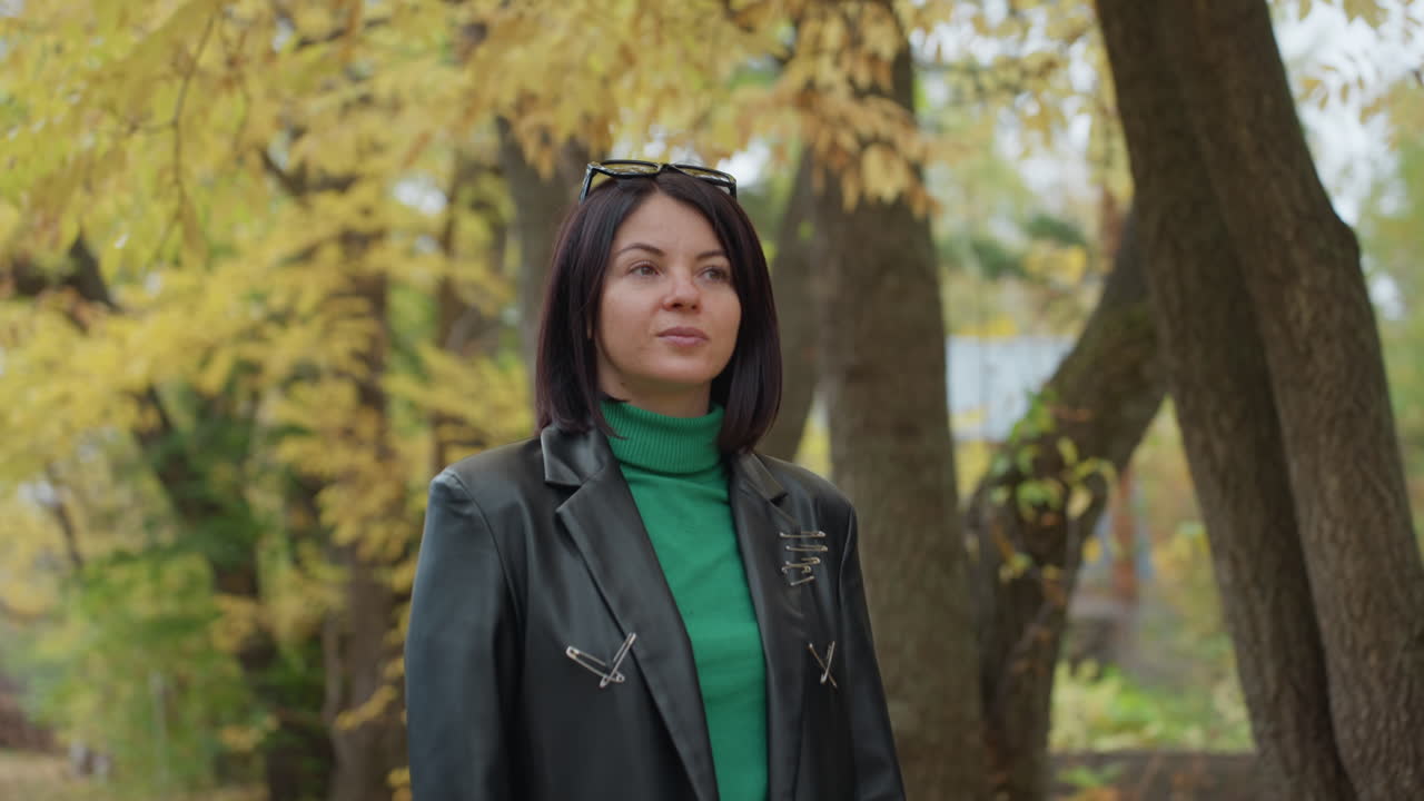 black woman pausing beneath golden canopy wearing leather jacket and green turtleneck, confident gaze and poised profile, autumn park backdrop with textured tree bark and drifting leaves, closeup