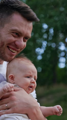 bearded man wearing white t-shirt holding a baby. loving dad waving his son and kissing kid's head. blurred nature backdrop. vertical video