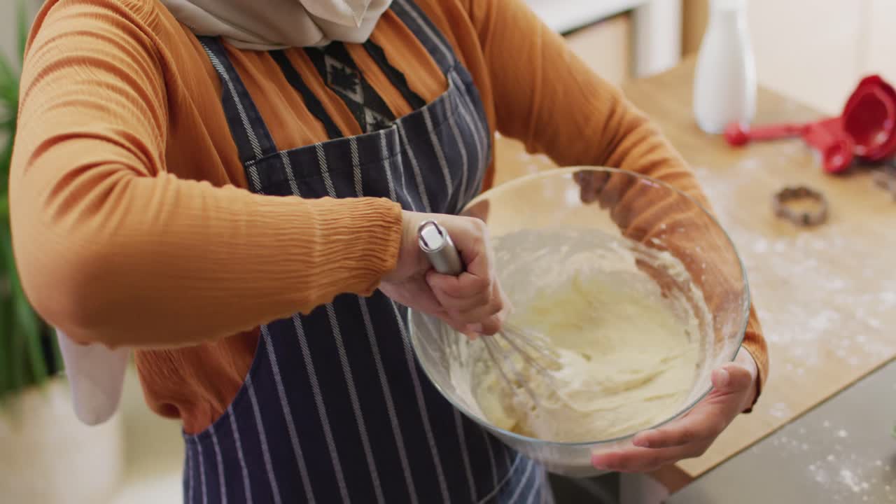 Video seorang wanita birasial berhijab yang ceria sedang memanggang di dapur, mengaduk adonan kue sambil memegang mangkuk adonan