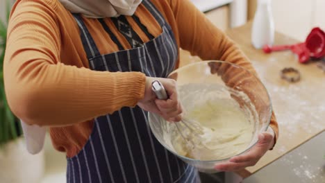 Video-of-happy-biracial-woman-in-hijab-baking-in-kitchen,-mixing-cake-batter-holding-mixing-bowl