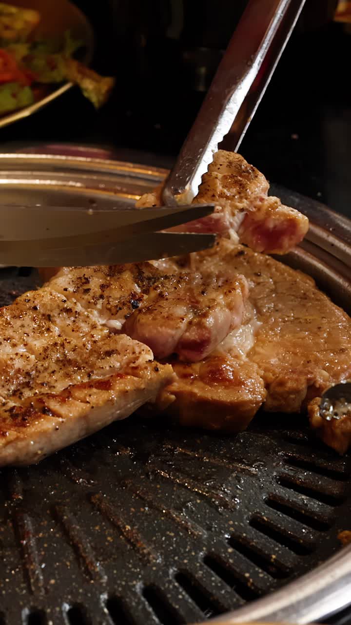 Hånd bruger tang til at skære grillet svinekød på bordplade koreansk barbecue under varm indendørs belysning