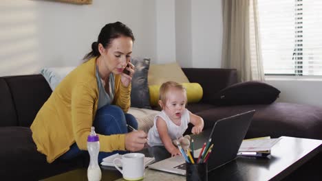 Caucasian-mother-holding-her-baby-taking-notes-and-talking-on-smartphone-while-working-from-home