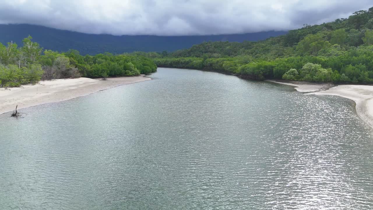 Drone glides above winding mangrove river, sandy banks, and lush rainforest under dramatic cloudy sky