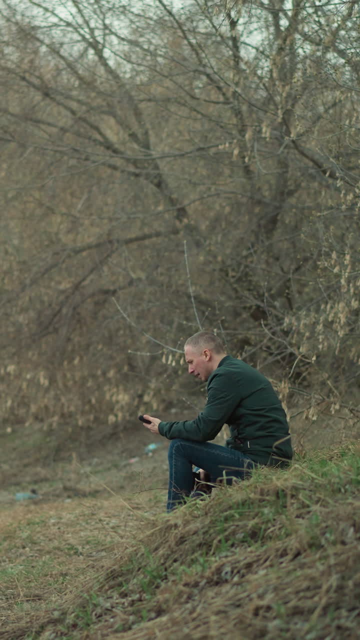A close view of a man in a green jacket sitting alone in a secluded, wooded area, seemingly deep in thought or contemplation, holding a cell phone and piston