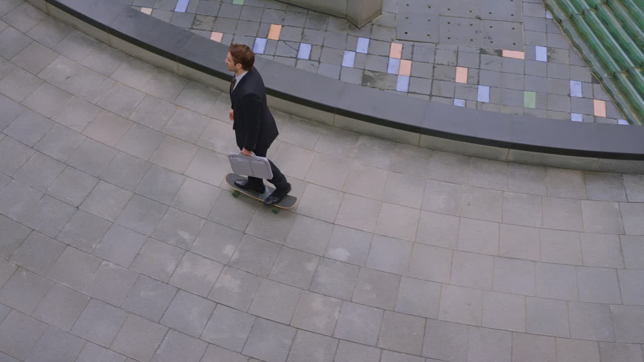 Campo lungo dal grandangolo di un giovane uomo d'affari in giacca che va sullo skateboard in slow motion portando una borsa per laptop attraverso una piazza urbana