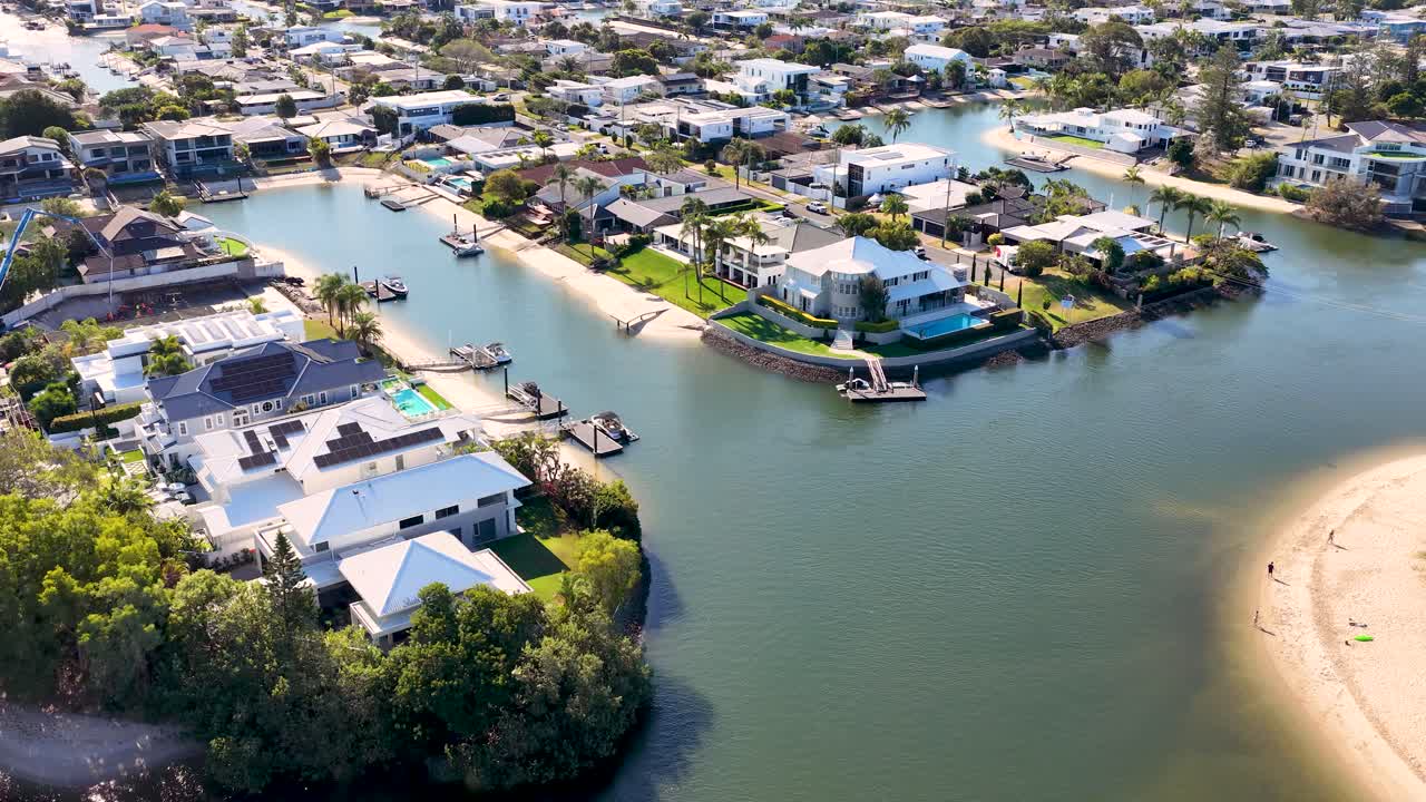 Drone glides above river, luxury houses, and sandy banks under bright daylight in Gold Coast