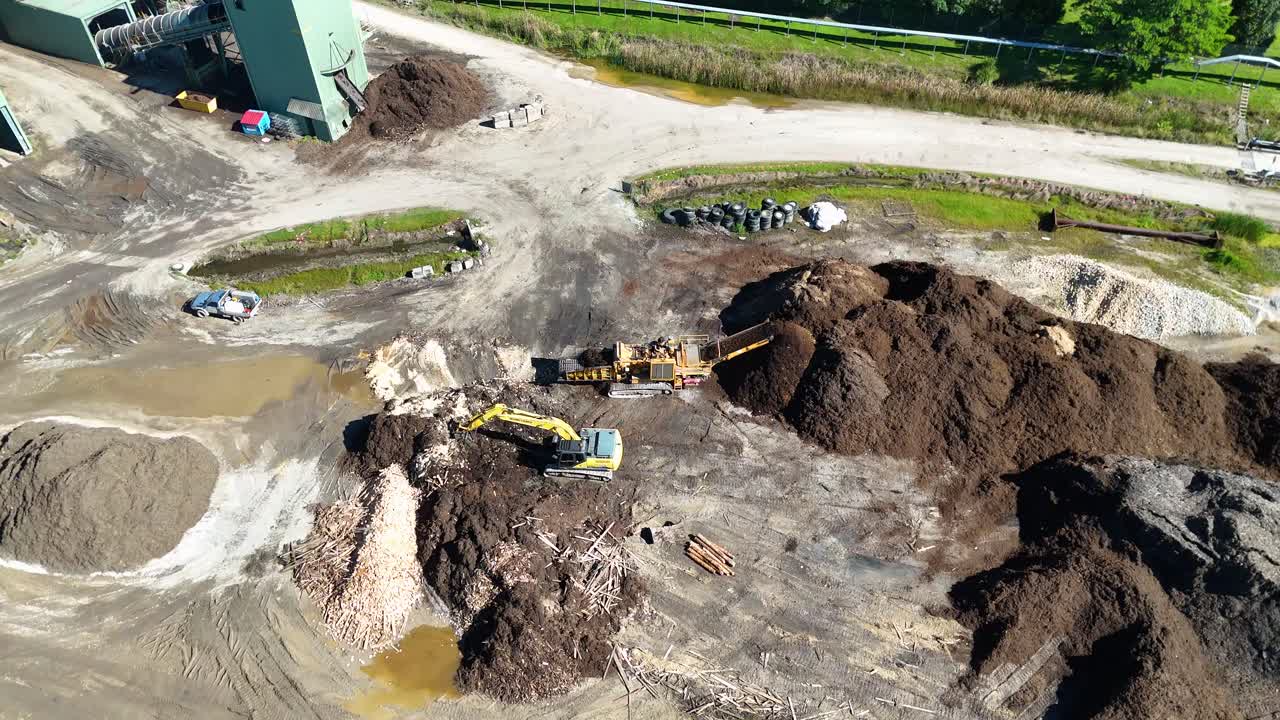 Drone captures machinery moving soil and mulch in a sunny industrial site. Dynamic aerial perspective highlights the scale and activity