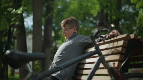 young boy stretches on wooden park bench, glancing back at his bicycle parked nearby, the scene is set in a peaceful, tree-lined park, with lush greenery, residential building in the background