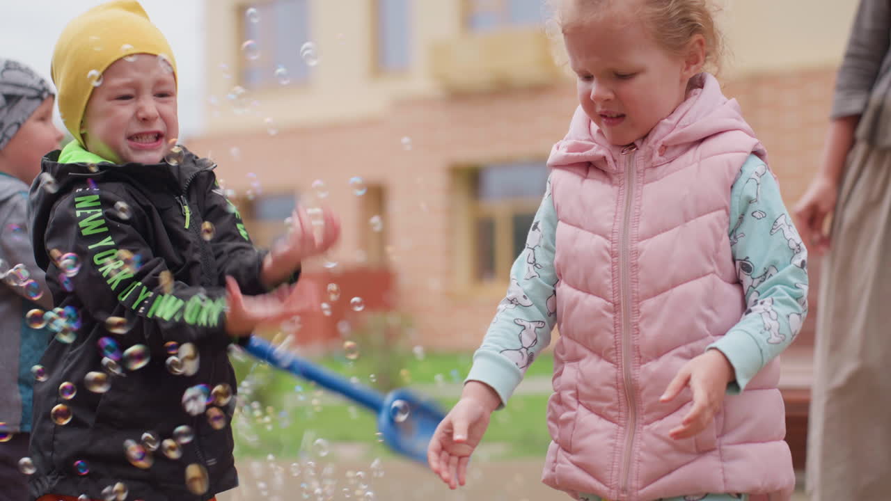 Young boy holds blue shovel while bubbles rise around him, girl in pink vest smiles watching nearby, adult stands close on courtyard scene with bench, lively outdoor moment filled with laughter