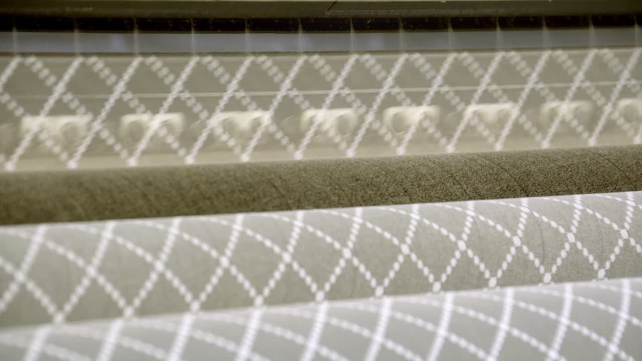 Detail vzorované látky a textilních rolí na výrobní lince, geometricky prošitý materiál pohybující se pod válečky průmyslového stroje v továrně na výrobu čalounění a bytového textilu.