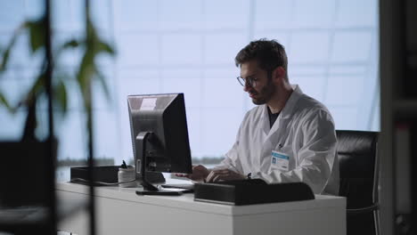 Tilt-down-shot-of-male-doctor-in-lab-coat-and-glasses-typing-on-laptop-while-working-at-desk-in-medical-office