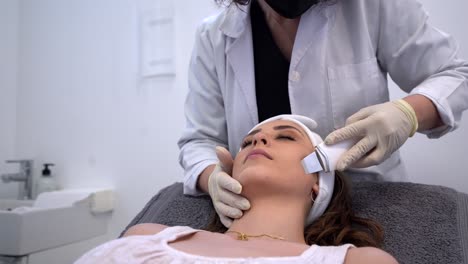 crop beautician doing ultrasound face cleaning for woman in clinic