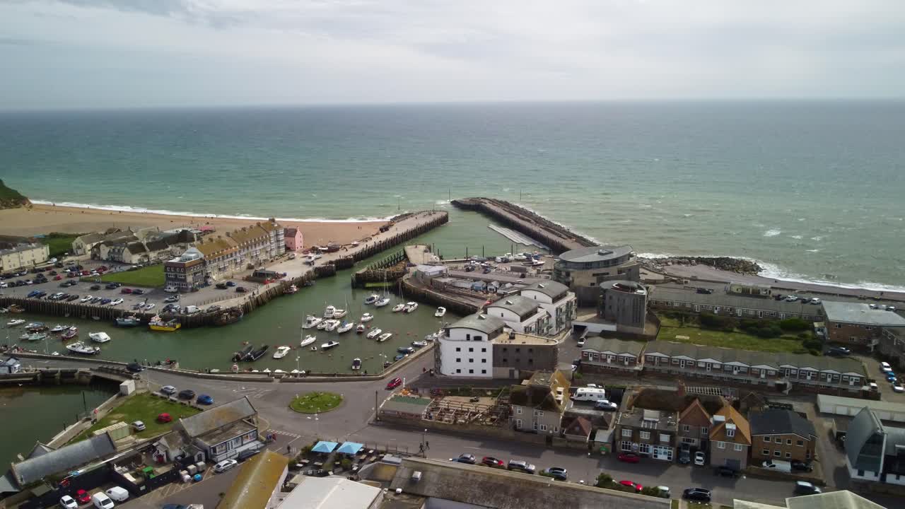 Aerial footage over West Bay, Dorset harbour. Orbiting around the harbour with the ocean behind. Wide view capturing most of the town, buildings, homes, business, roads with car driving on and boats.