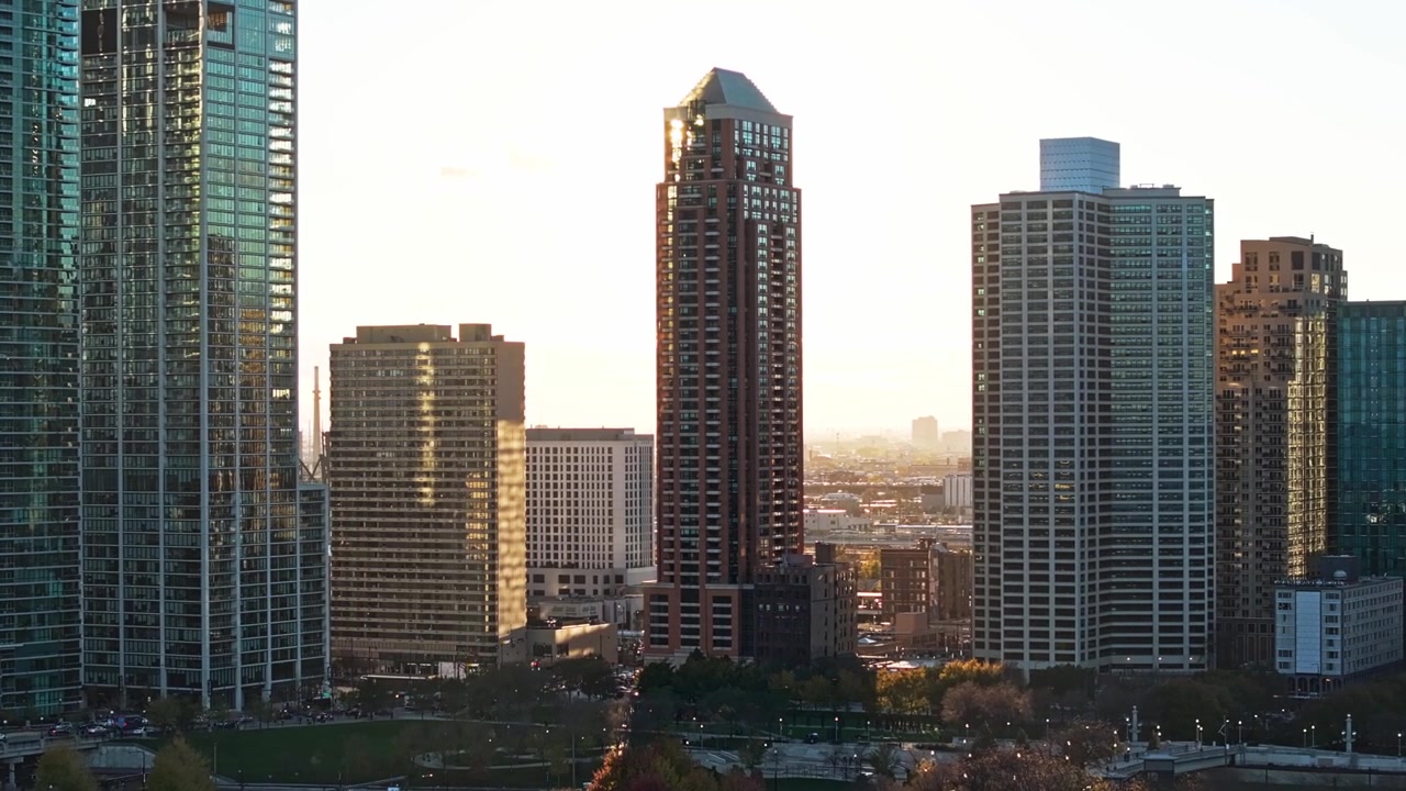 Avvicinamento aereo a un edificio retroilluminato e luminoso nello skyline del centro di Chicago, con alti grattacieli e riflessi durante l'ora d'oro.