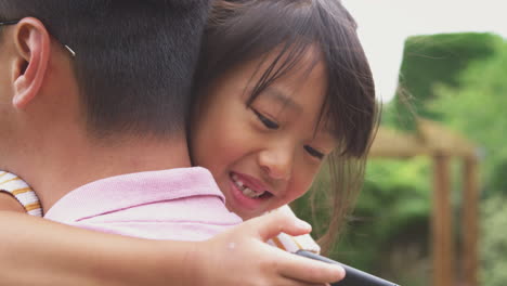 asian father cuddling girl in garden as girl looks over his shoulder at mobile phone