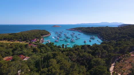 Yachten-In-Der-Bucht-Nahe-Der-Küstenstadt-Kroatiens-Vor-Dem-Hintergrund-Des-Blauen-Himmels-Und-Des-Blauen-Transparenten-Wassers,-Grüner-üppiger-Bäume-Und-Häuser-Mit-Roten-Dächern