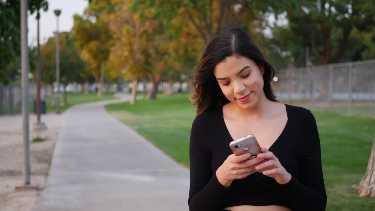 bella joven hispana independiente que se ve feliz mientras revisa su teléfono en busca de mensajes de texto o notificaciones de redes sociales en un paseo por el parque