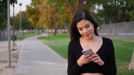 Schöne-Junge,-Unabhängige-Hispanische-Frau-Sieht-Glücklich-Aus,-Während-Sie-Bei-Einem-Spaziergang-Durch-Den-Park-Ihr-Telefon-Auf-Textnachrichten-Oder-Social-Media-Benachrichtigungen-überprüft