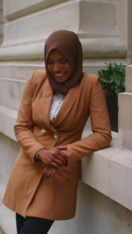 vertical video portrait of smiling muslim businesswoman wearing hijab and modern business suit standing outside city office buildings 10