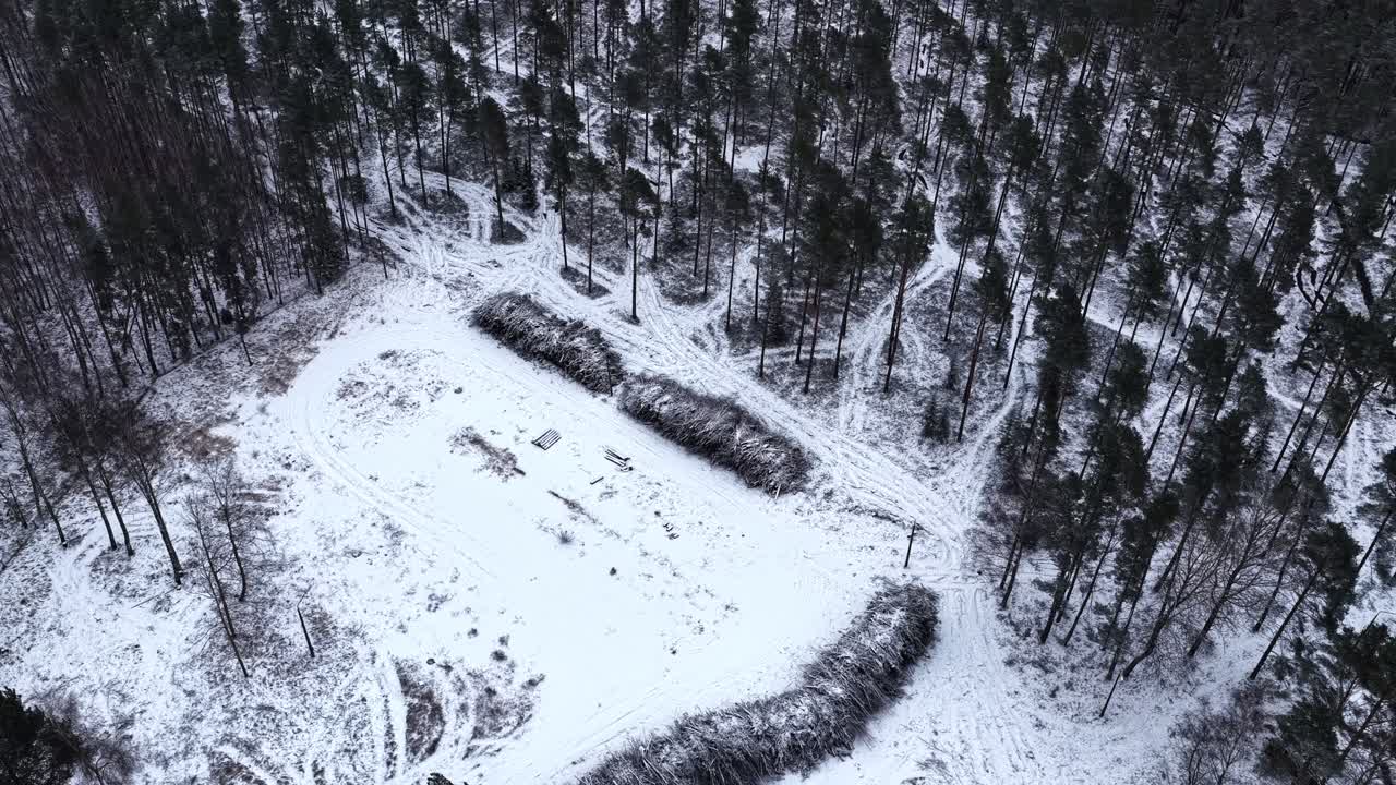 Ripresa aerea con drone che esegue lentamente una panoramica e sale sopra una radura innevata, rivelando cataste di tronchi, tracce di veicoli e alberi circostanti coperti di neve.