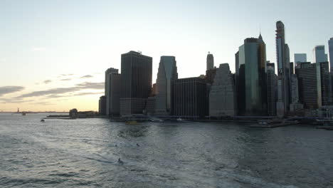 Wide-angle-pull-back-shot-of-Lower-Manhattan,-New-York-City-with-jet-skis-and-boats-on-the-East-River-at-dusk-on-a-clear-summer-evening