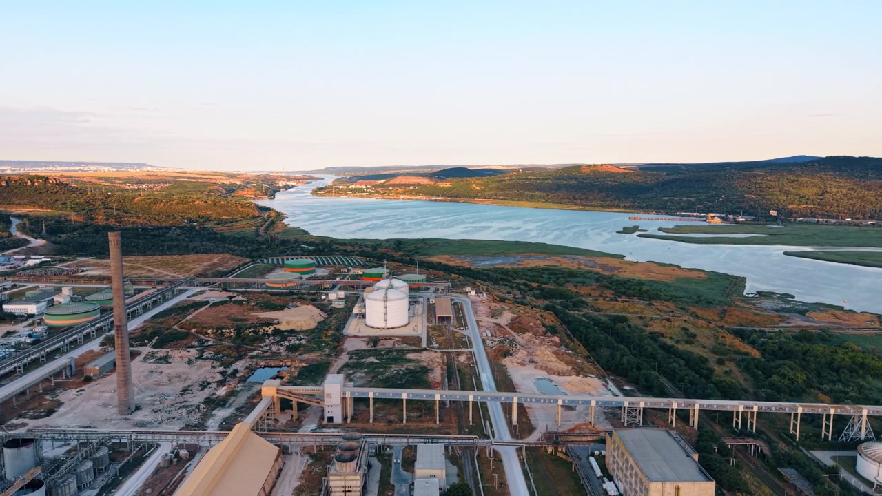 Vista aerea di un impianto industriale lungo un ampio fiume. Edifici di produzione e serbatoi di stoccaggio si trovano accanto a un grande specchio d'acqua circondato da verdi colline