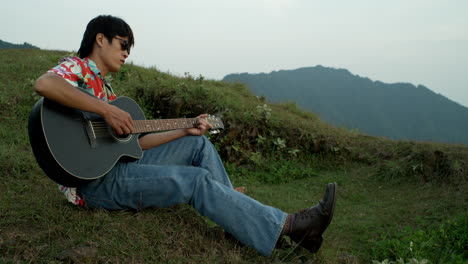 asian male musician plays guitar watching the sky at mountain green landscape wearing hawaiian shirt, vintage jeans and boots, countryside hills