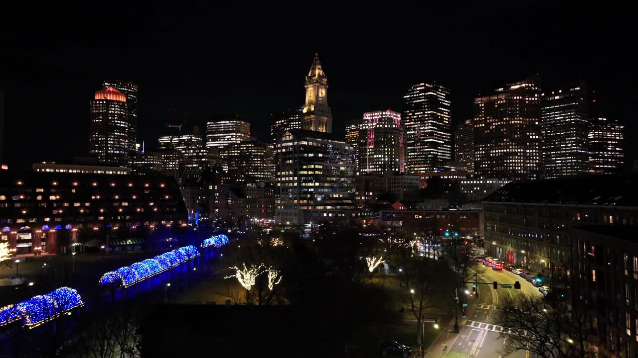Illuminated Custom House Tower and Boston skyline at night. Vibrant city streets and tree-lit park in the foreground. Urban aerial.