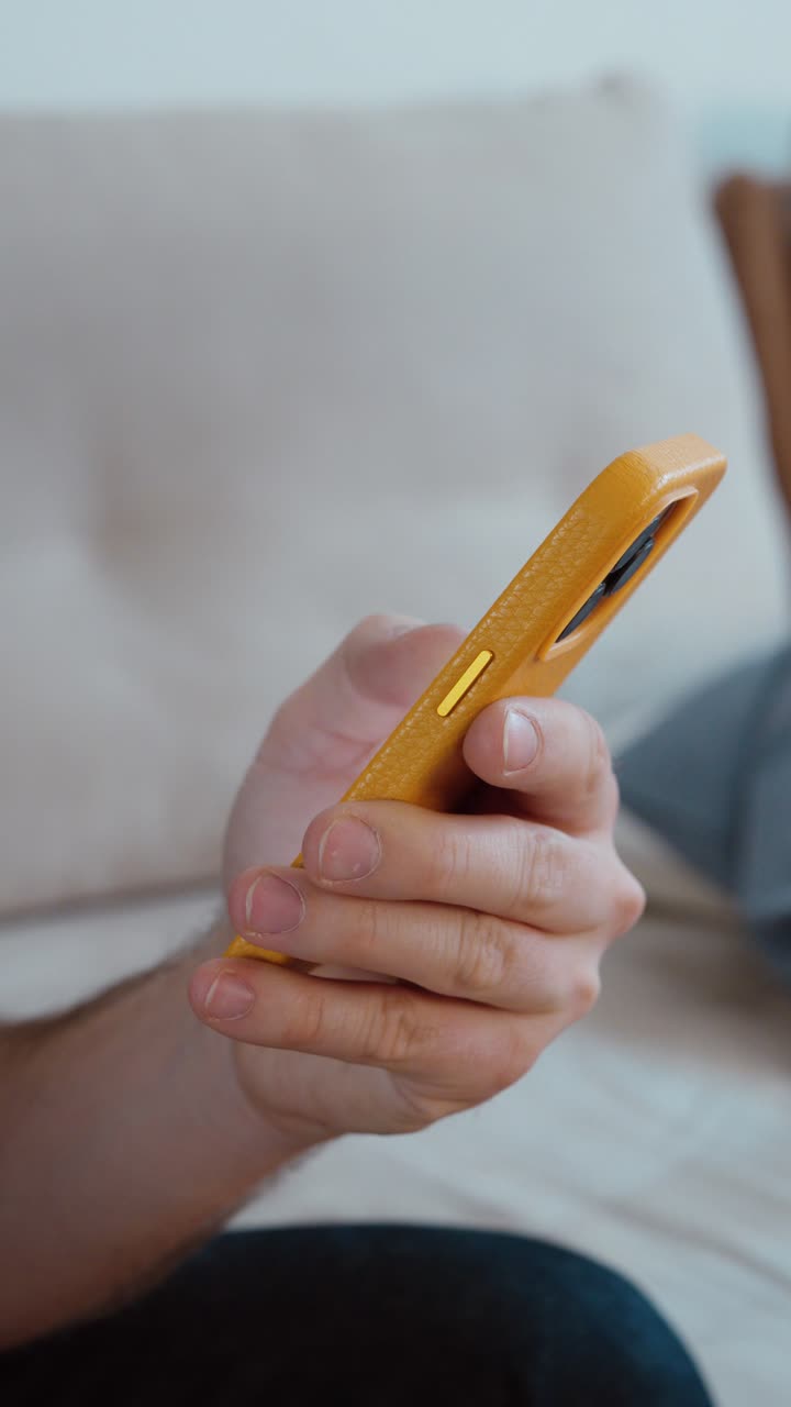 Primo piano verticale di un uomo che tiene uno smartphone con uno sfondo di un divano morbido, che mostra l'uso casuale del cellulare, lo stile di vita domestico moderno e l'interazione digitale quotidiana in un ambiente interno rilassato
