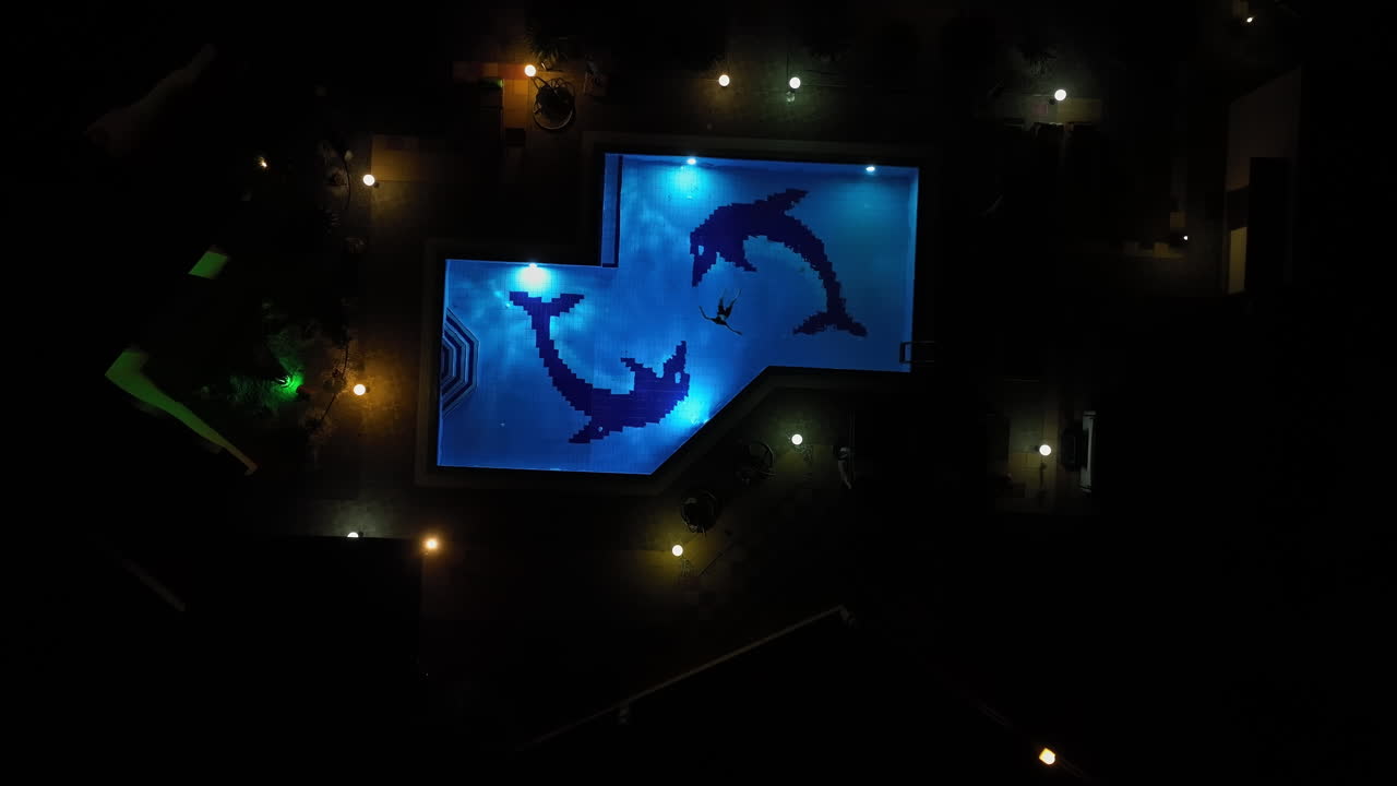 Top down drone shot above a boy swimming in a pool decorated with dolphins, night