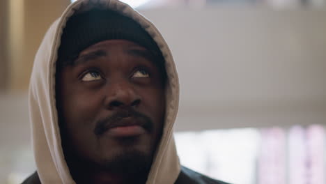 young black chap in beige hoodie and black beanie gazing upward inside sunlit modern interior space with blurred windows and reflective metal elements conveying curiosity and calm introspection