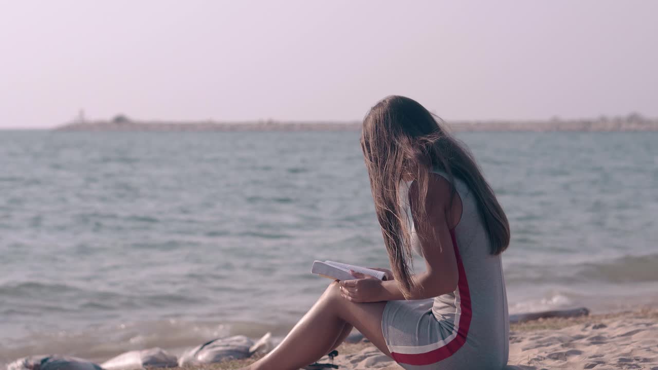 긴 머리카락의 갈색 여성이 바다 모래 해변에서 책을 읽습니다.