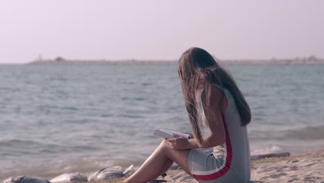una morena de cabello largo lee un libro en la playa de arena del océano