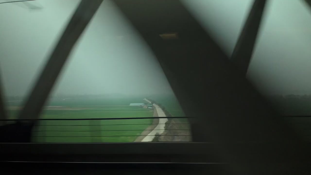 Landscape passes rapidly outside train window while crossing bridge structure in rural China. Motion blur captures speed of bullet train traveling through green fields under overcast sky.