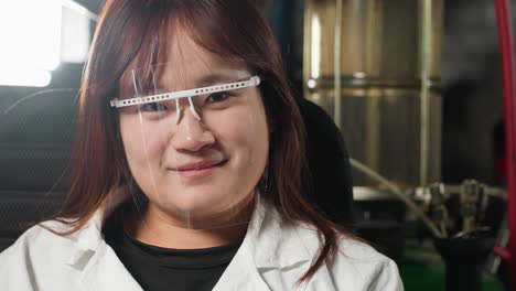 student wearing protective face shield blinks eyes with warm smile in mechanical workshop, background features industrial tools, bright lighting, and machinery