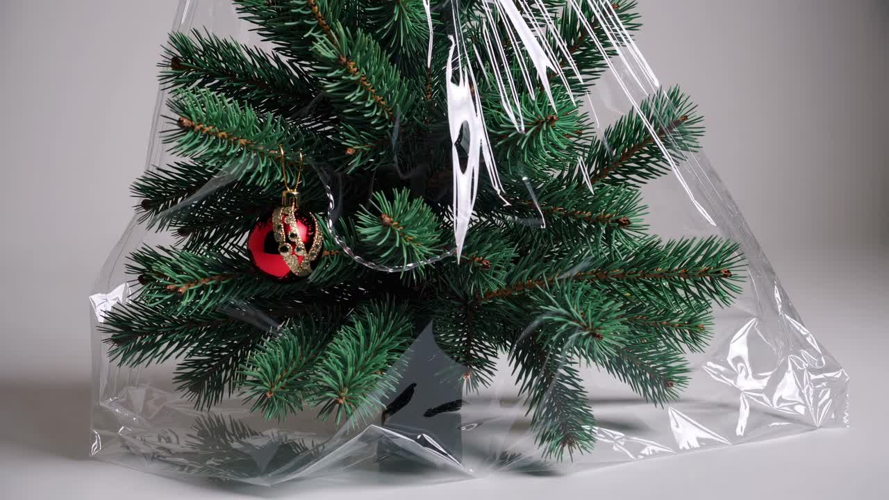 Small decorated christmas tree wrapped in transparent plastic film, symbolizing consumerism, packaging and environmental issues related to plastic use during holidays