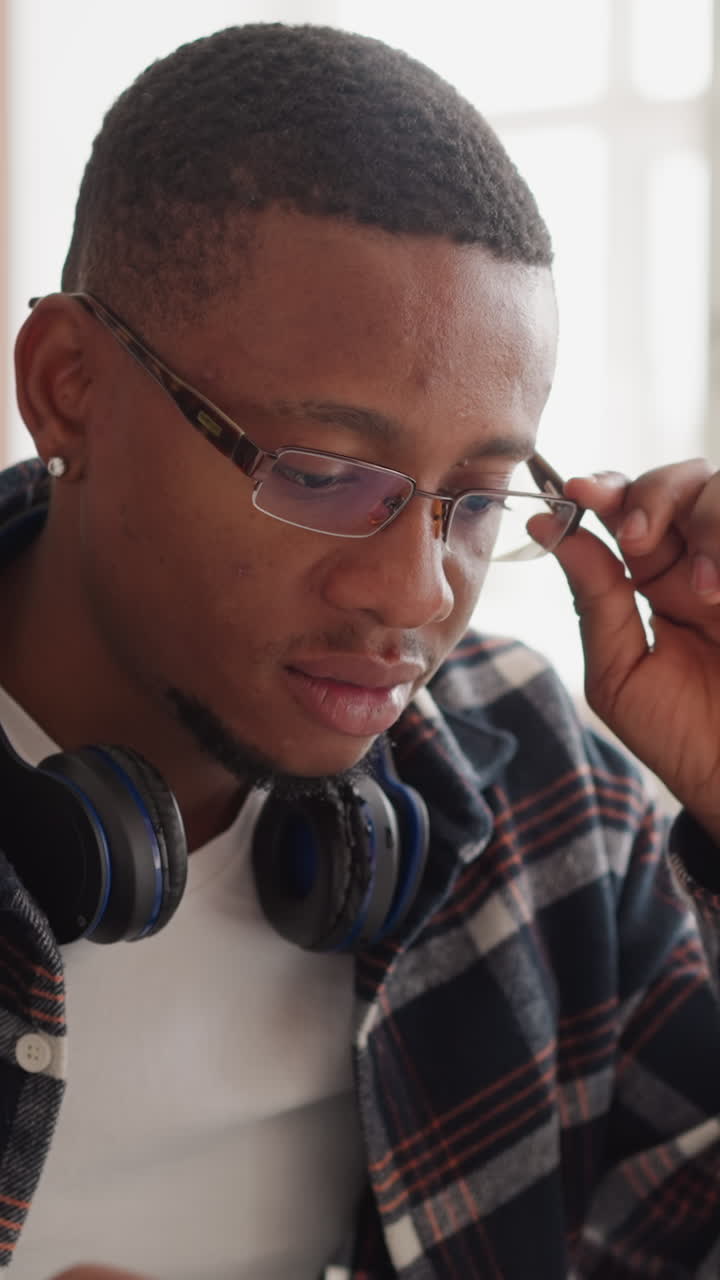 estudiante con gafas usa la computadora en la biblioteca. hombre afroamericano con auriculares se quita las gafas mirando atentamente a la pantalla. preparación para exámenes en la universidad