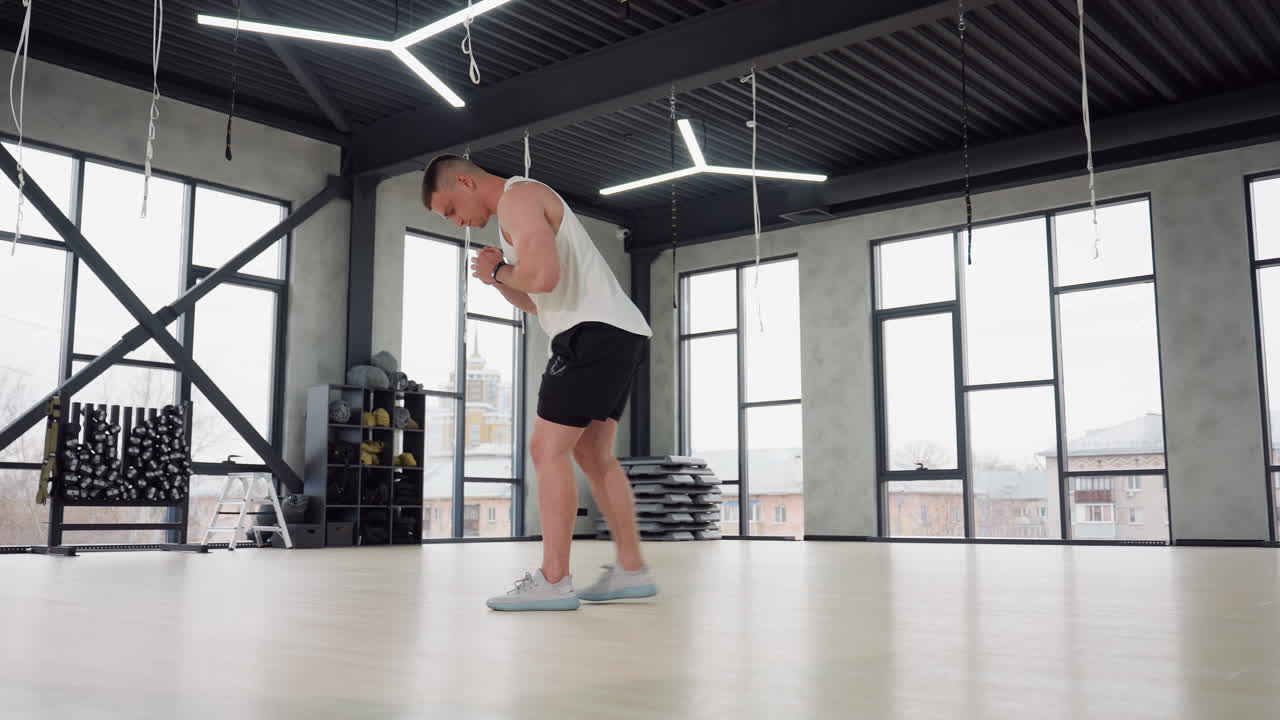 Athlete stretching legs with side lunge in spacious gym room, wearing white tank top and black shorts, large windows and hanging straps overhead, rubber floor beneath, conveying warmup, and focus